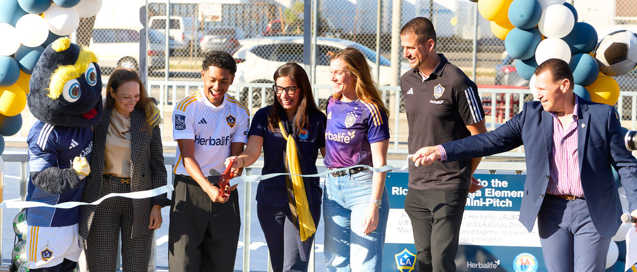 Ribbon-cutting ceremony at Ricardo Lizarraga Elementary School’s new mini-pitch in South Central Los Angeles, featuring LA Galaxy mascot, Herbalife representatives, and soccer-themed decorations with balloons and signage.